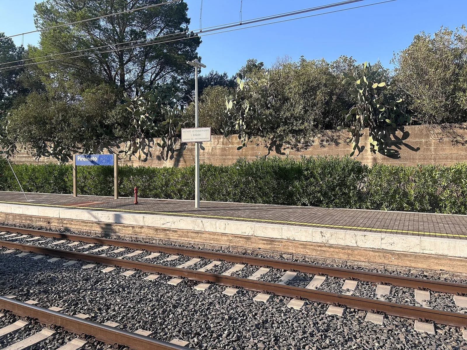 Banchina della stazione ferroviaria di Piraineto con il cartello del nome, binari e fichi d'India